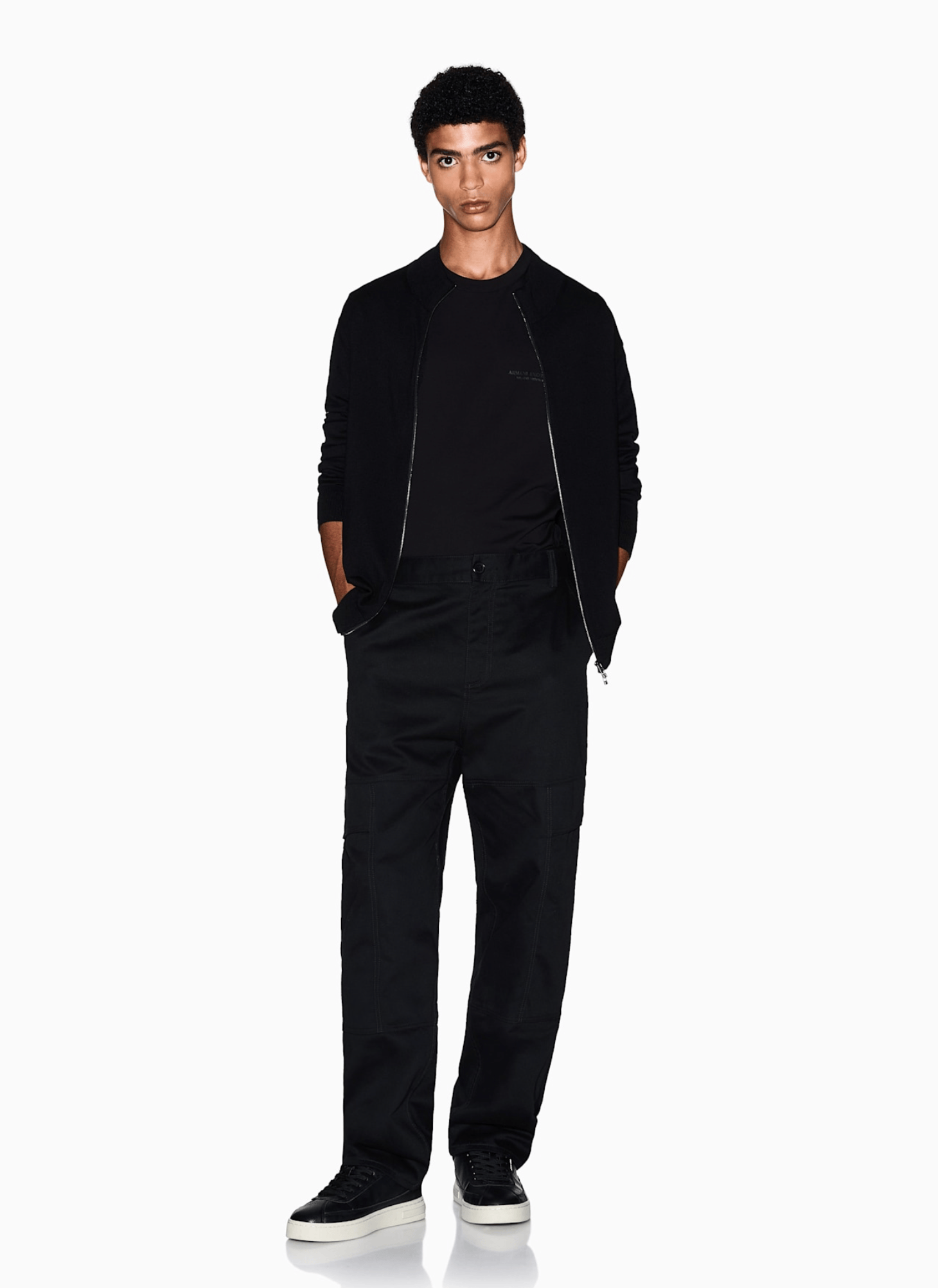 A male model wearing a black zip-up bomber jacket, black t-shirt, black cargo pants, and black sneakers with white soles, standing against a solid white studio background.