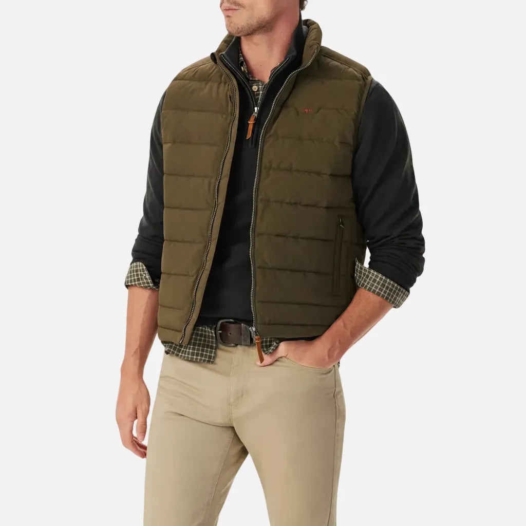 Man wearing a green quilted vest over a black shirt with plaid sleeves, beige pants, and a belt.