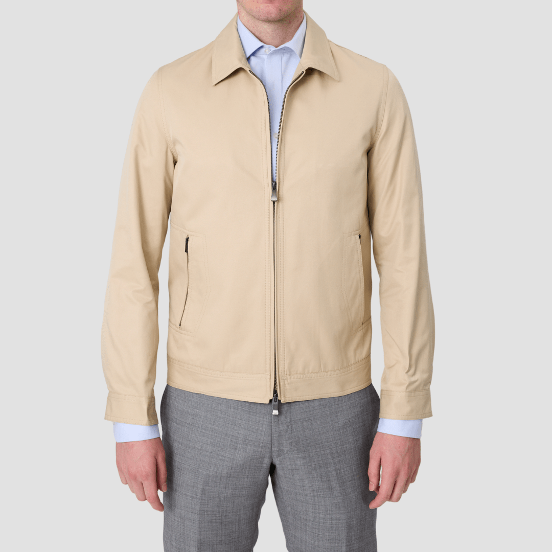 A man wearing a stone-colored zip front jacket with a regular collar, double zip pockets, snap waist adjusters, and snap button cuffs.