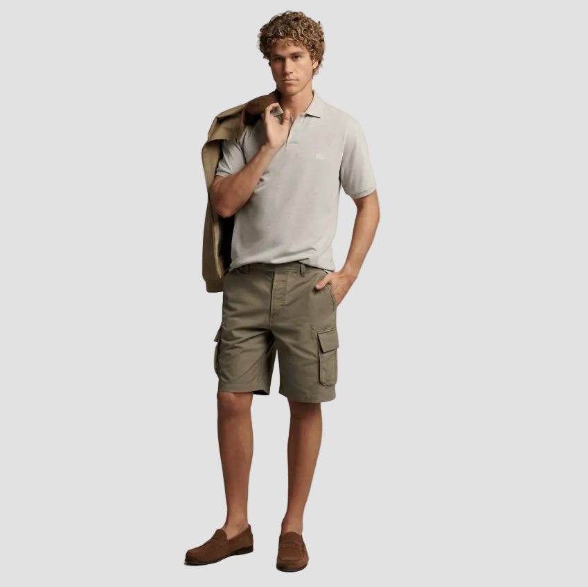 A male model stands against a plain beige studio background wearing a light gray short-sleeve polo shirt, olive cargo shorts, and brown suede loafers, with a tan jacket draped over his shoulder.