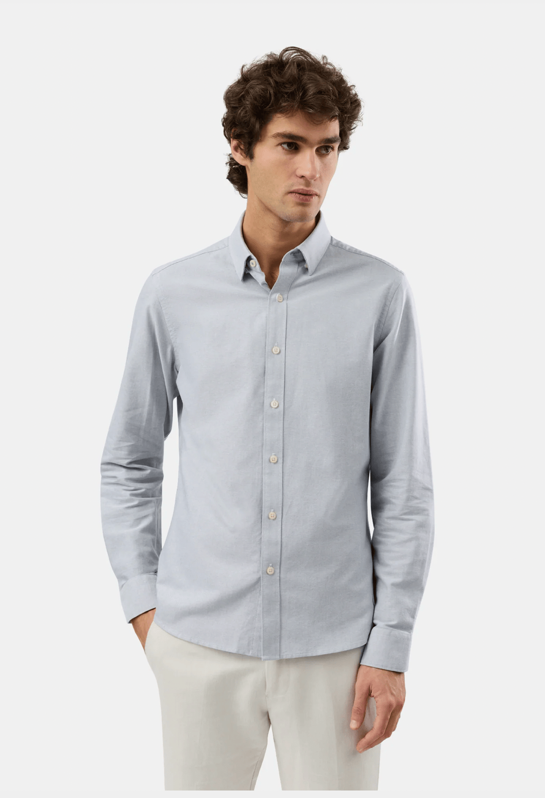 A man wearing a light blue long-sleeve button-down shirt and beige trousers, standing against a plain light gray studio background.