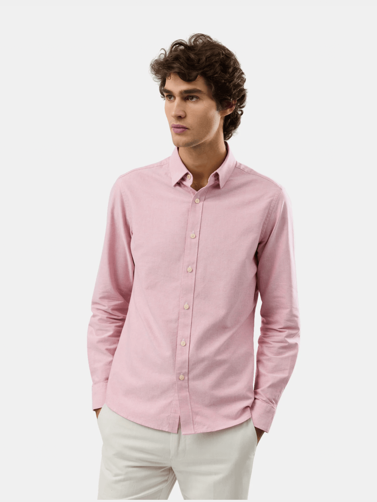 A man wearing a light pink long-sleeve button-down shirt and off-white trousers, standing against a plain light gray studio background.