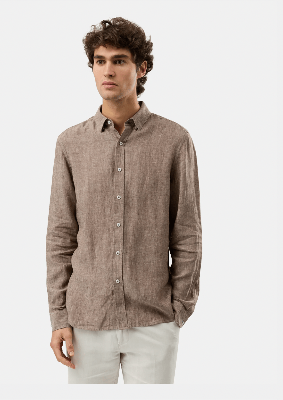 A man wearing a light brown linen button-down shirt and off-white trousers, standing against a solid light gray background.