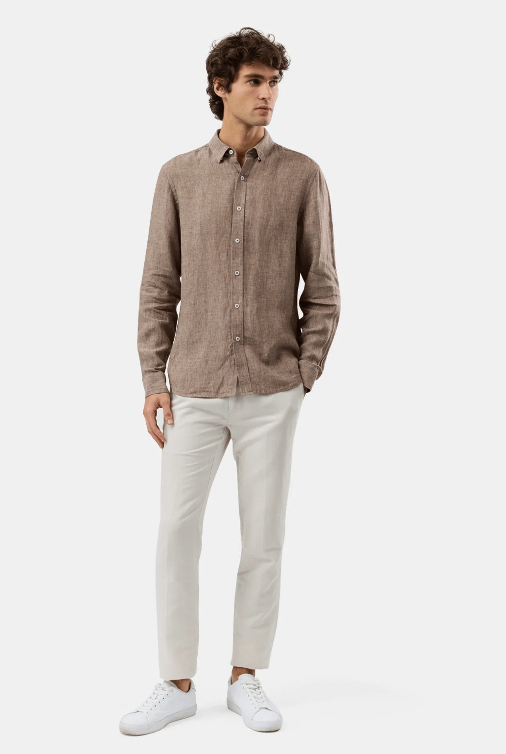 A man modeling a light brown linen button-down shirt, paired with off-white trousers and white sneakers, against a plain white studio background.