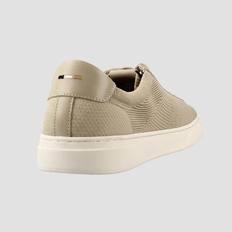 A beige slip-on sneaker with a textured knit upper, smooth leather heel tab, and a thick white platform sole, shown from a three-quarters rear angle on a white background.