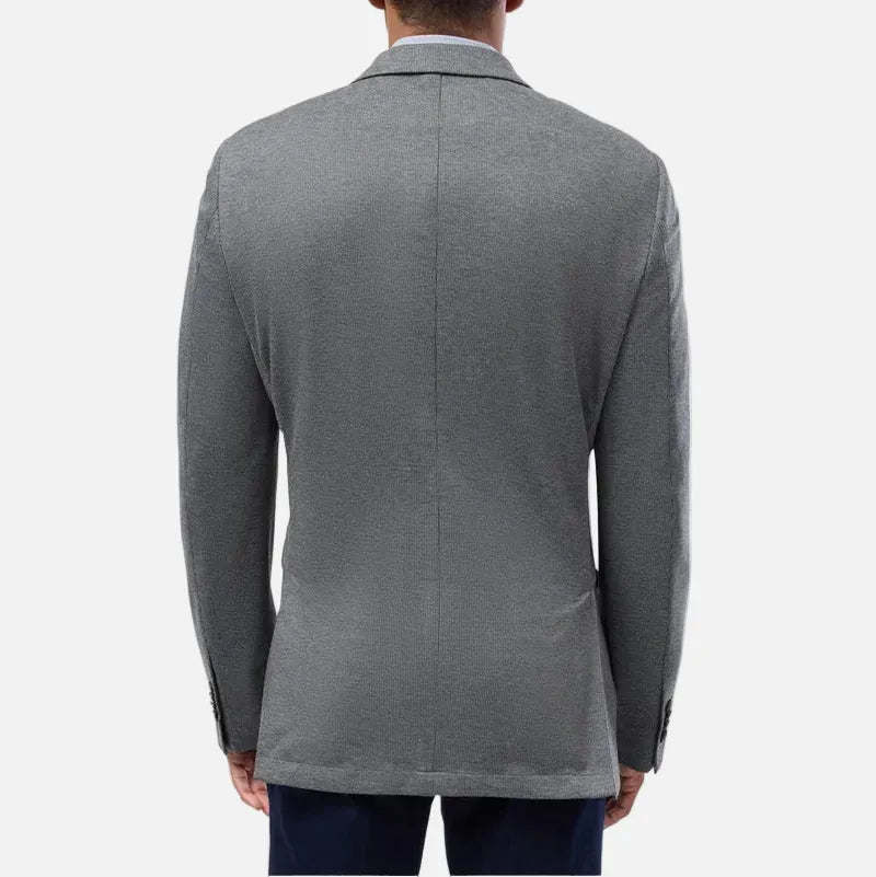 A man viewed from the back wearing a gray knit blazer over a white collared shirt and navy trousers, set against a plain light gray studio background.
