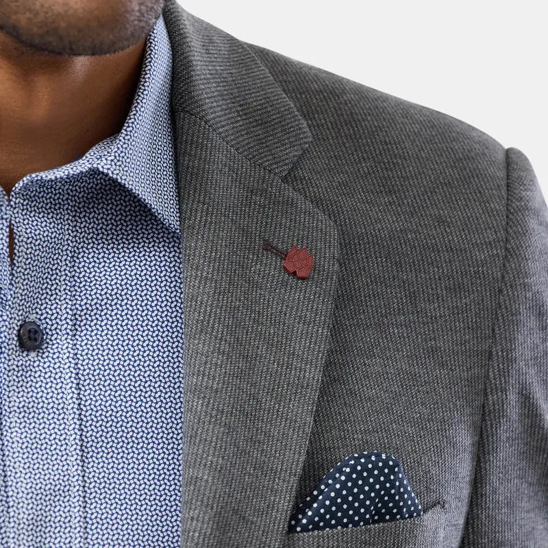 Close-up of a man wearing a gray textured blazer over a light blue patterned dress shirt, with a small brown lapel pin and a blue-and-white polka dot pocket square.