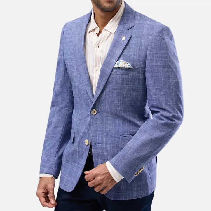 A man wearing a blue plaid blazer over a cream striped dress shirt and dark trousers, standing against a light gray studio background.