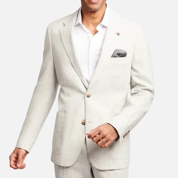 A smiling man wearing a light beige linen suit jacket with a white dress shirt and a patterned pocket square, posed against a solid light gray background.