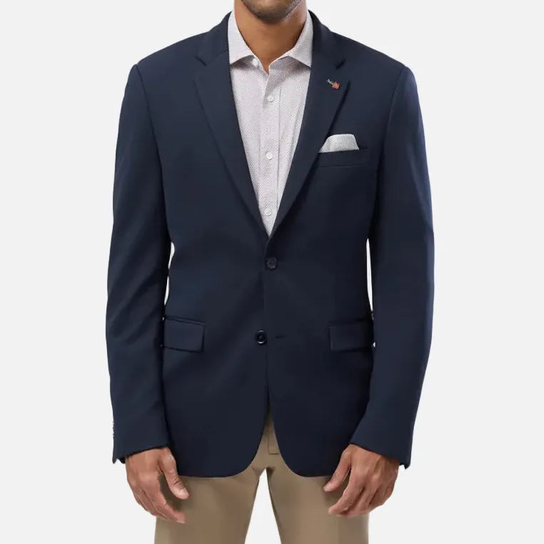 A man wearing a navy blue blazer over a light-colored dress shirt and khaki pants, standing against a solid light gray background.