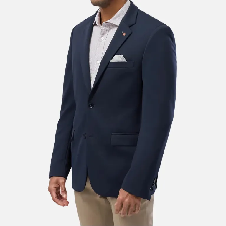A man wearing a navy blue blazer over a light-colored collared shirt and khaki pants, standing against a light gray studio background.