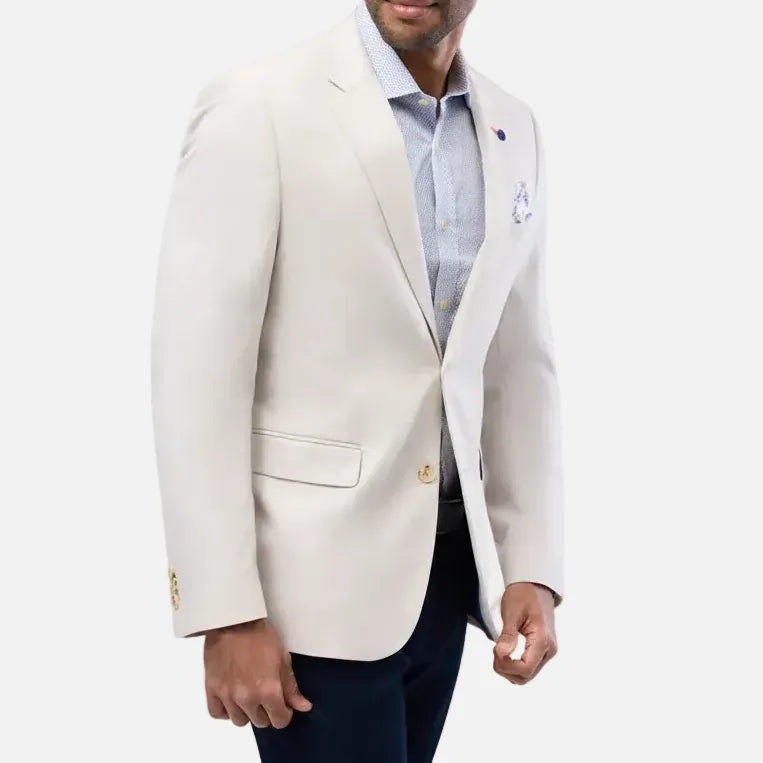 A man wearing a light cream-colored blazer over a light blue dress shirt and dark trousers, standing against a solid light gray background.