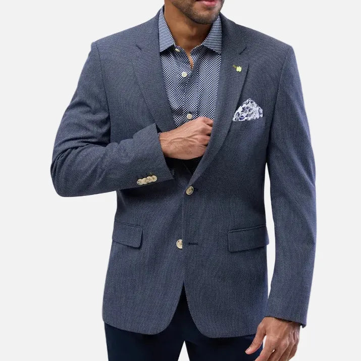 A man wearing a navy blue pinstriped blazer over a matching navy patterned dress shirt, with a white pocket square and dark trousers, photographed against a light gray studio background.