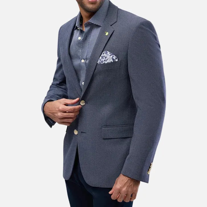 A man wearing a navy blue textured blazer over a patterned blue shirt, with a matching patterned pocket square, posed against a light gray background.