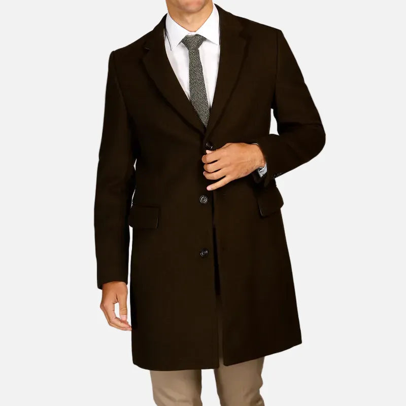 A man wearing a dark brown mid-length overcoat over a white dress shirt and gray tie, standing against a light gray studio background.