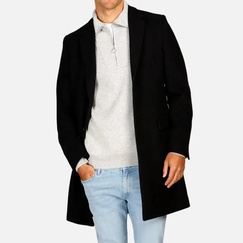 A man modeling a black mid-length overcoat worn open over a light gray polo sweater and light blue jeans, photographed against a plain light gray studio background.
