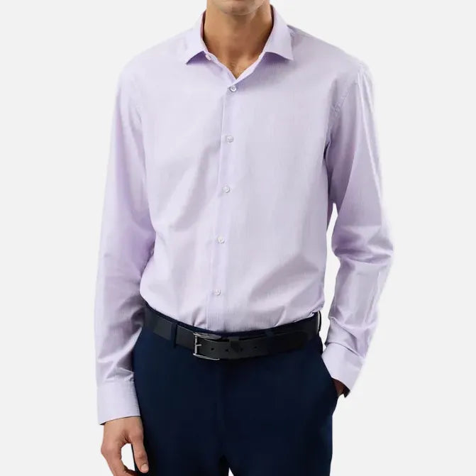 A man wearing a light lavender long-sleeve dress shirt, dark navy trousers, and a black belt, standing against a plain light gray studio background.