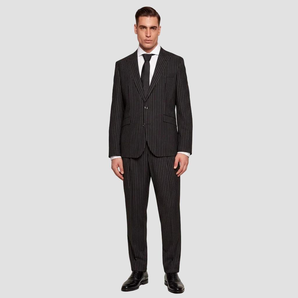 A man wearing a black pinstripe suit with a white dress shirt and a black tie, standing against a light off-white background.