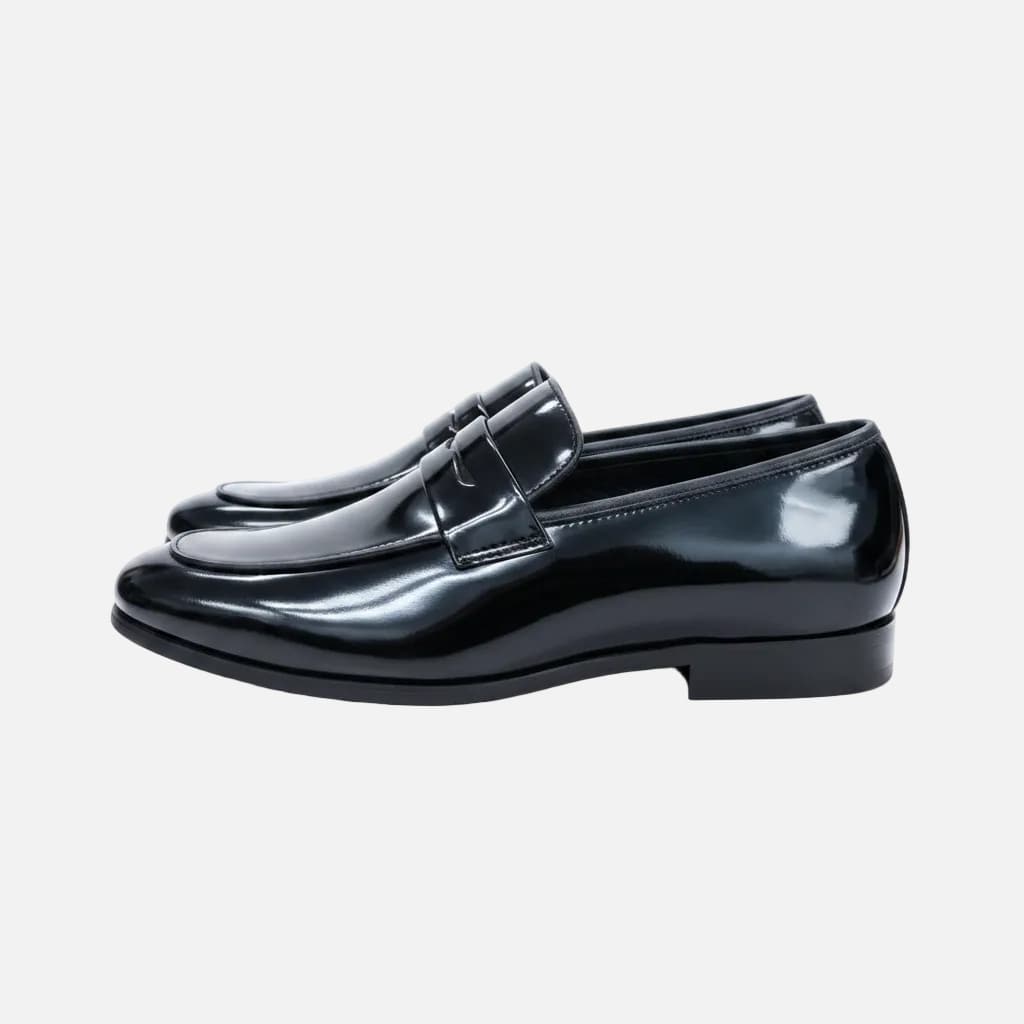 A pair of shiny black leather penny loafers with a low block heel, displayed on a light wood table with a clothing store background.