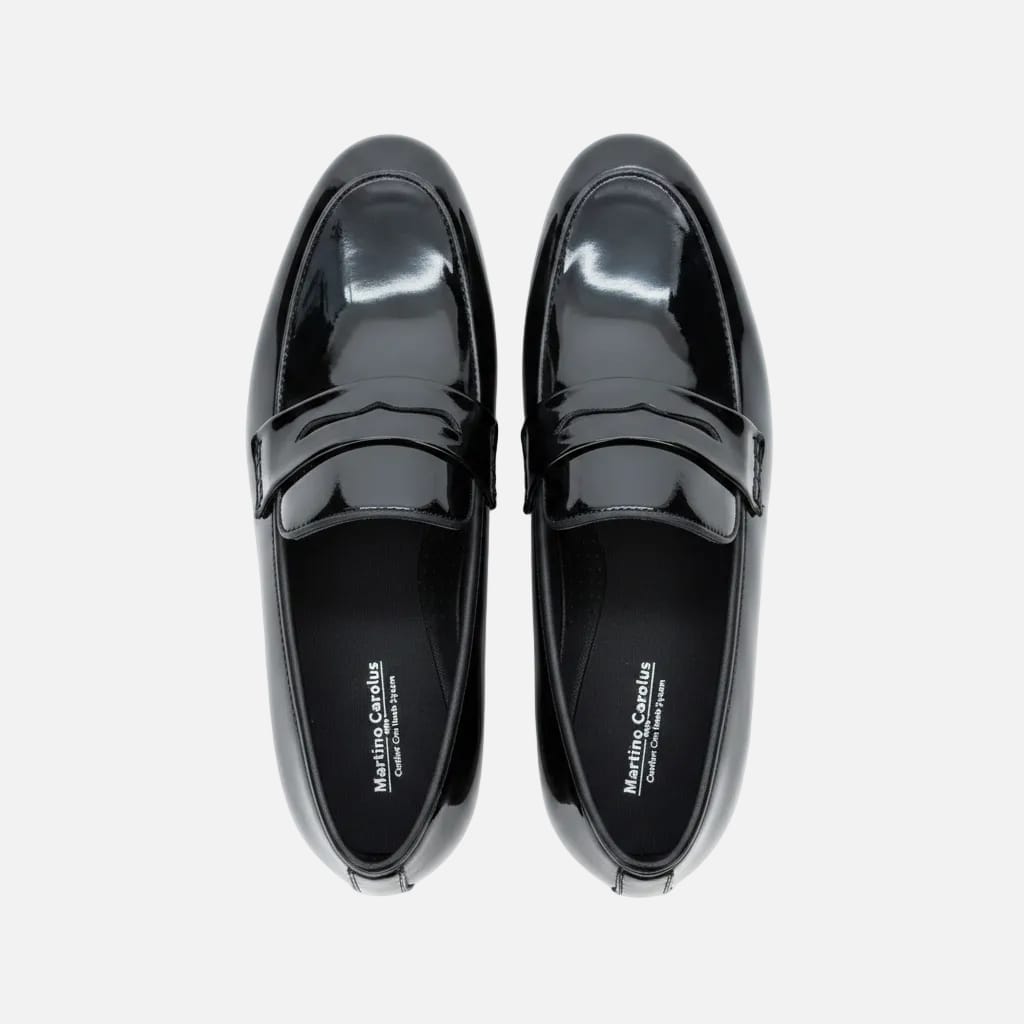 A pair of glossy black patent leather penny loafers with double straps, laid flat on a light brown wooden surface. The brand name 'Matteo Croce' is visible on the insole.