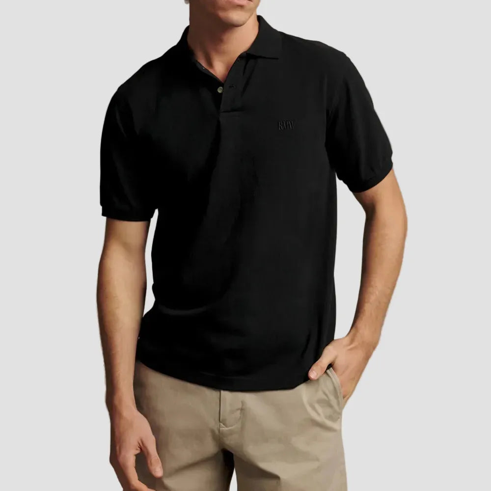 A man wearing a black short-sleeve polo shirt and beige trousers, standing against a plain beige studio background.