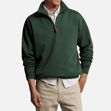 A man modeling a dark green quarter-zip pullover sweater over a white collared shirt, paired with beige trousers and brown shoes, standing against a plain beige studio background.