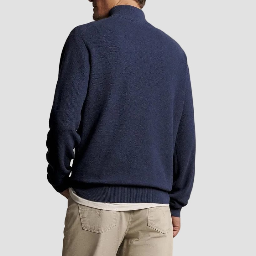 A man shown from the back wearing a navy blue mock-neck knit sweater and beige trousers, standing against a solid warm gray studio background.
