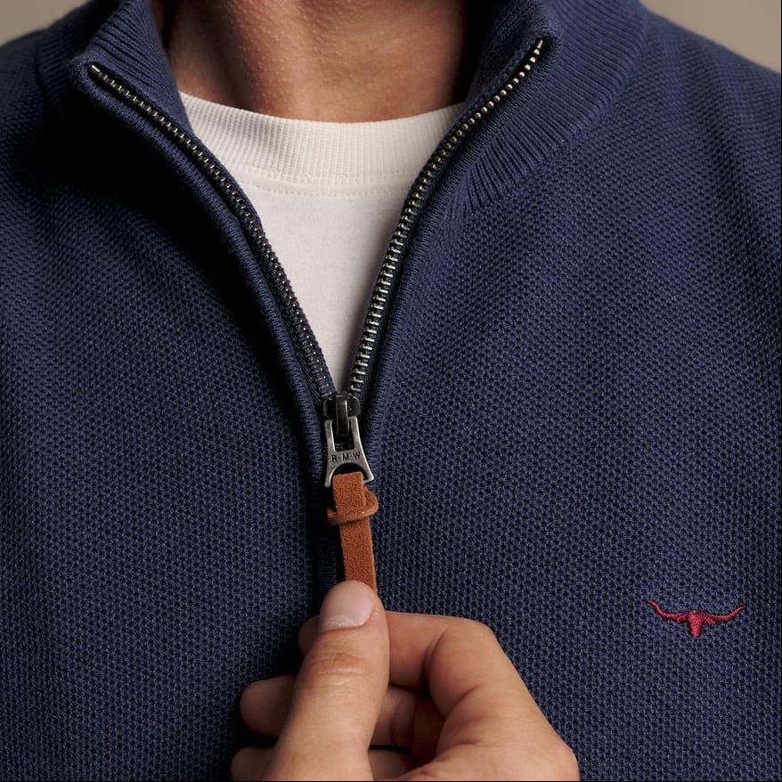 Close-up of a man wearing a navy blue half-zip knit sweater with a brown leather zipper pull, over a white crew-neck t-shirt. A small red embroidered logo is visible on the left chest.