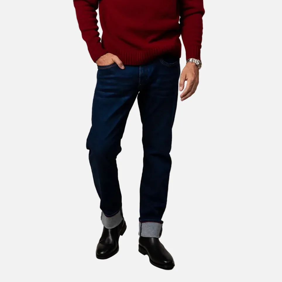 A man wearing a deep burgundy knit crewneck sweater, dark blue jeans, and black leather shoes, standing against a solid beige studio background.