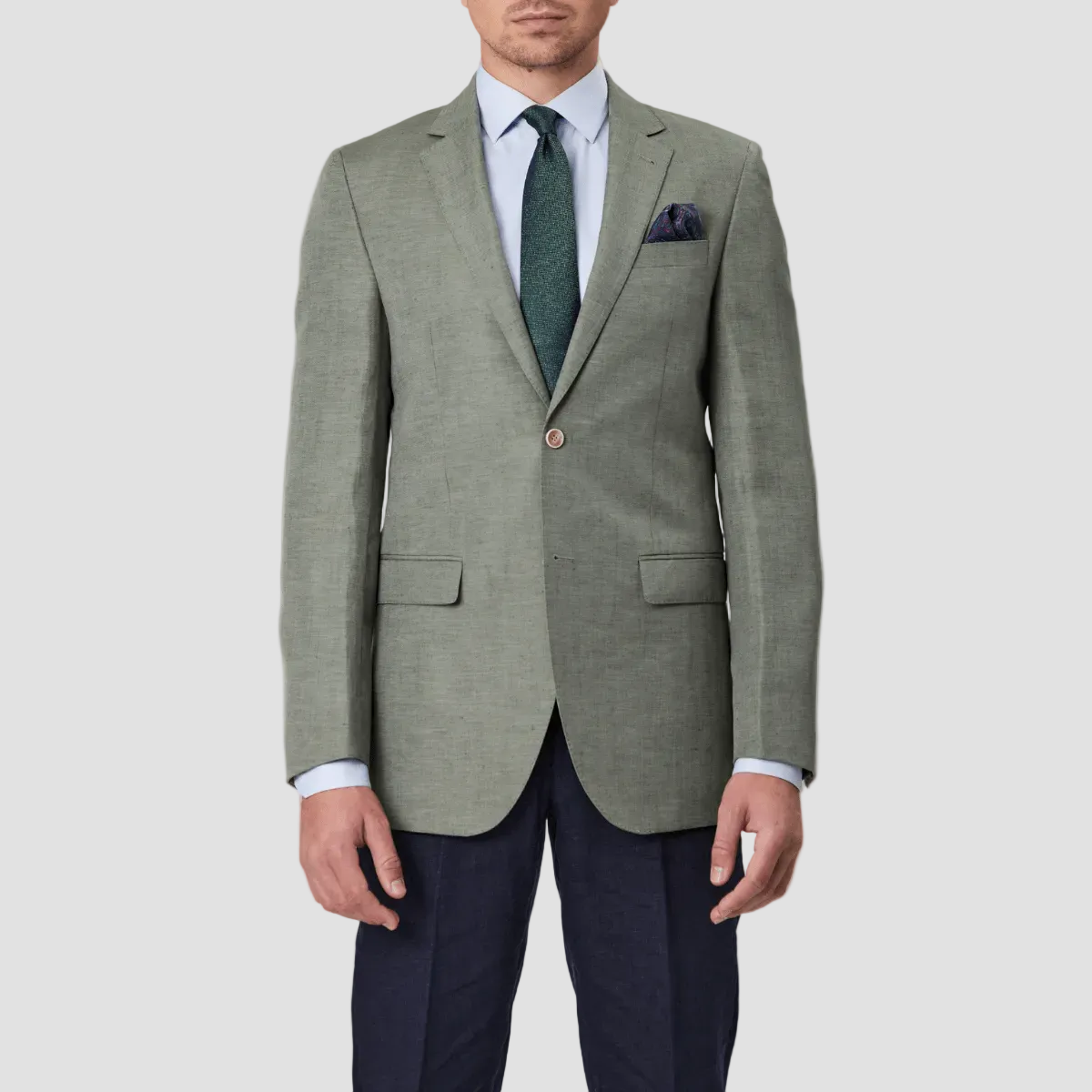 A man wearing a green slim fit sports jacket with notch lapel, double button closure, and welt chest pocket, paired with navy pants, dark green tie and pocket square.
