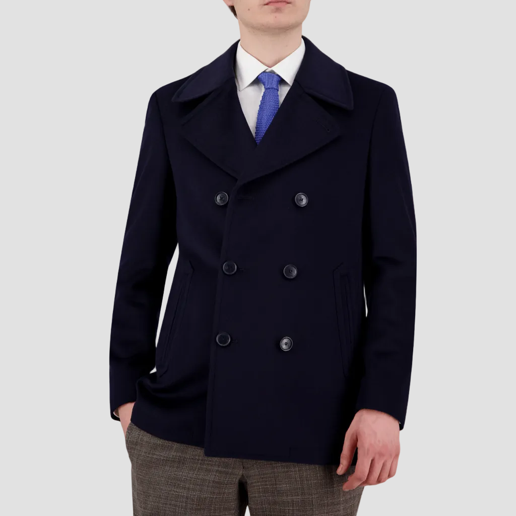 A man wearing a navy double-breasted pea coat with button sleeves and two side pockets, paired with a white shirt and a blue tie.