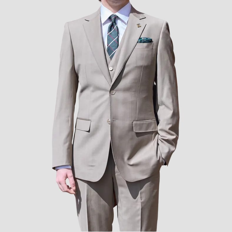 A man wearing a light beige two-piece suit, a white dress shirt, a blue and green striped tie, and sunglasses, standing against a solid light gray background.