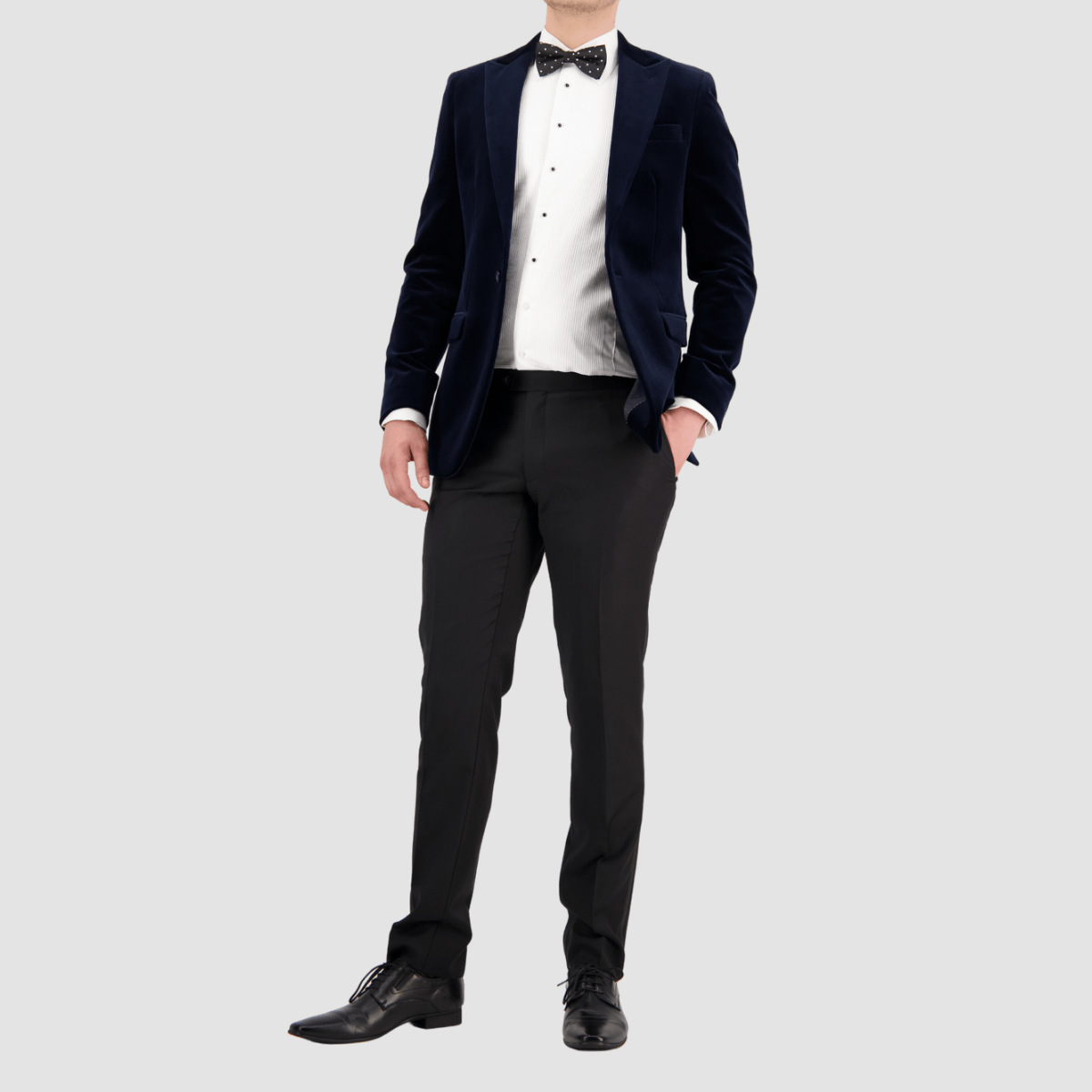A man wearing a navy velvet sports jacket with a slim fit and peak lapel by Savile Row, paired with a white shirt, black pants and a bow tie.