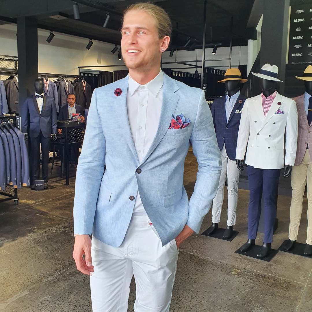 a man wears a light blue linen blazer over a white chino and white shirt, styled with a colour pocket-chief, he sands in a store with racks of suits and mannequins behind him 