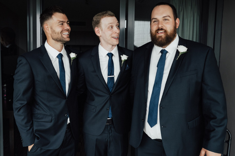 a groom and groomsmen wear dark coloured navy formal suits for a wedding celebration