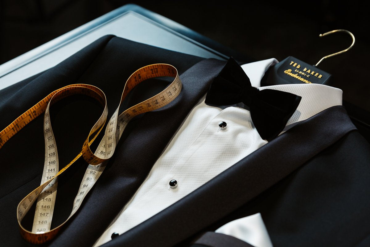 a mens black tuxedo by ted baker with a satin shawl collar, underneath is a white pin tucked shirt with contrasting black buttons