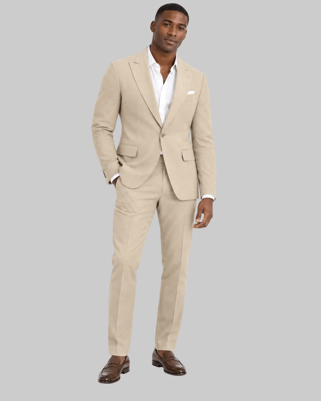 A man modeling a light beige linen suit with a white dress shirt and brown leather loafers, standing against a solid white background.