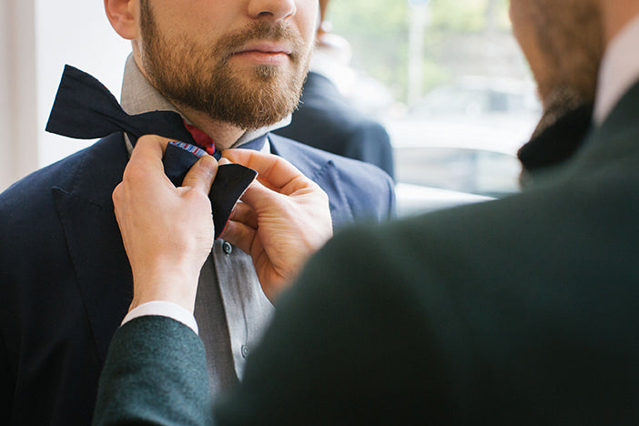 a groomsmen in a a navy blue formal suit gets his tie altered by the groom on the wedding day