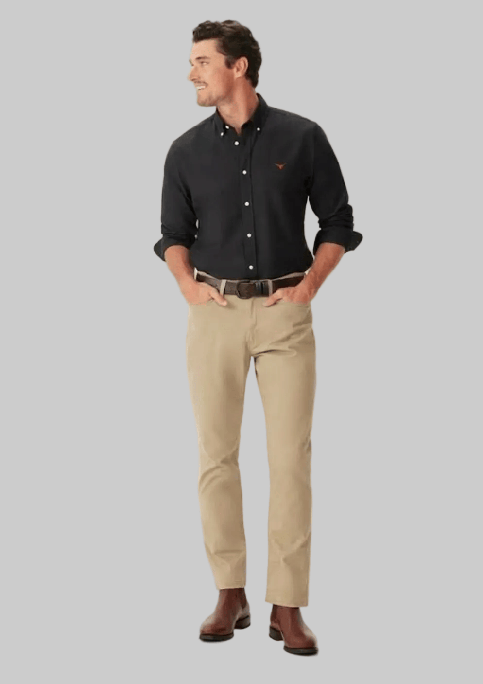 A man modeling a black long-sleeve button-down shirt with a small red embroidered logo on the chest, paired with khaki trousers and brown leather shoes, against a solid light beige background.