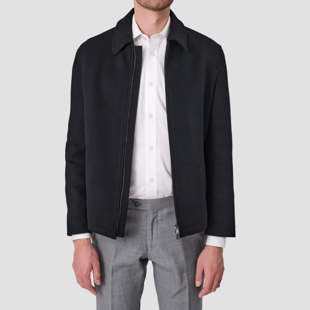 A man wearing a black zip front jacket by Savile Row with a regular collar over a white shirt and grey trousers.