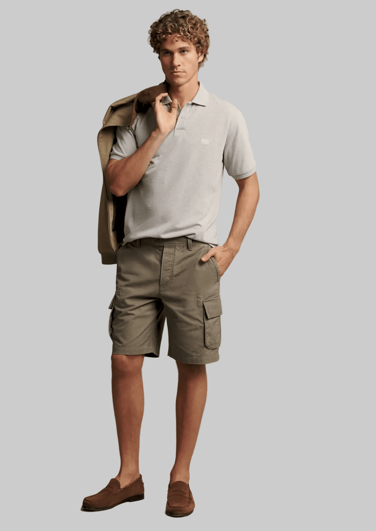 A male model stands against a plain beige studio background wearing a light gray short-sleeve polo shirt, olive cargo shorts, and brown suede loafers, with a tan jacket draped over his shoulder.