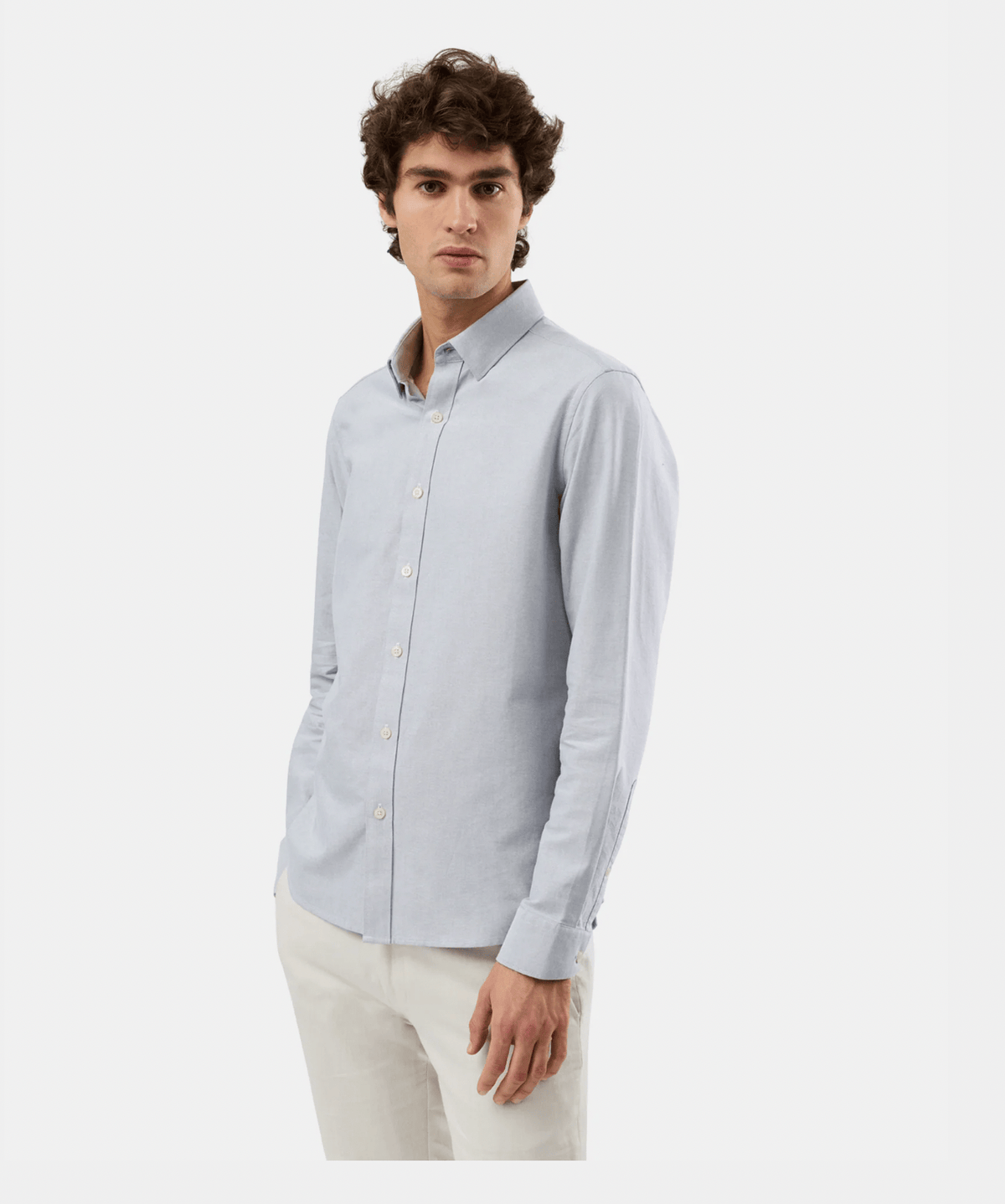A man wearing a light blue long-sleeve button-down shirt and off-white trousers, standing against a plain light gray studio background.
