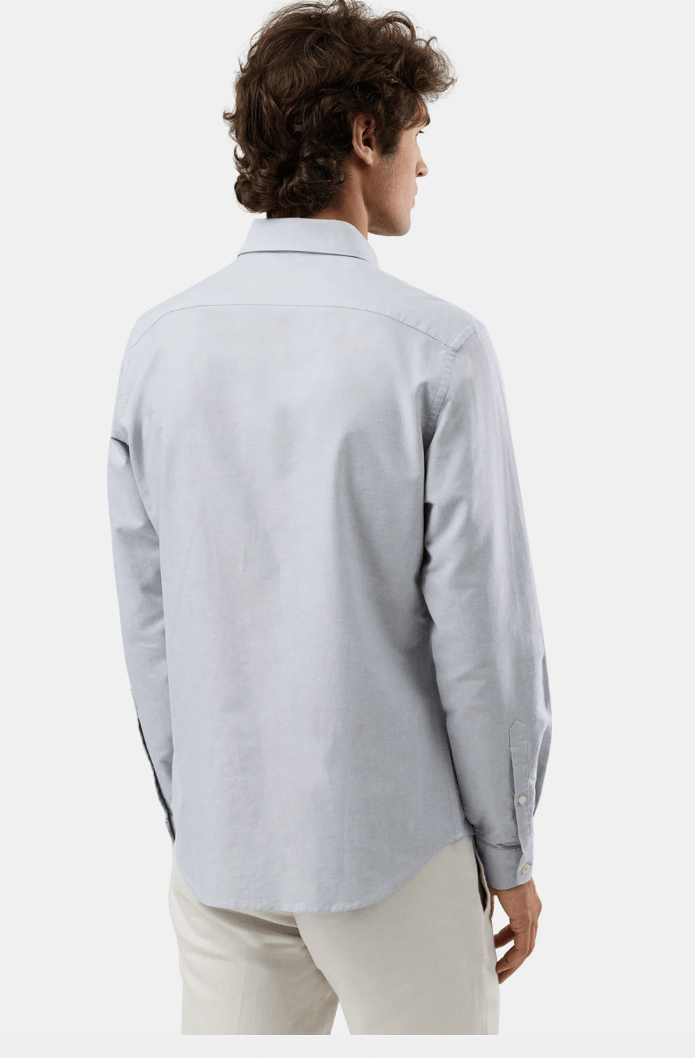 A man shown from the back wearing a light gray long-sleeve button-down shirt and off-white trousers, photographed against a plain light gray studio background.