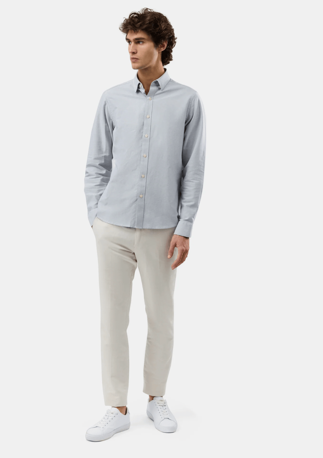 A man modeling a light blue long-sleeve button-down shirt with cream trousers and white sneakers, standing against a plain light gray studio background.