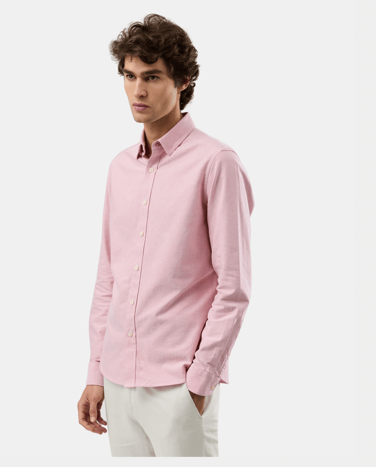 A man wearing a light pink long-sleeve button-down shirt and off-white trousers, photographed against a clean light gray studio background.