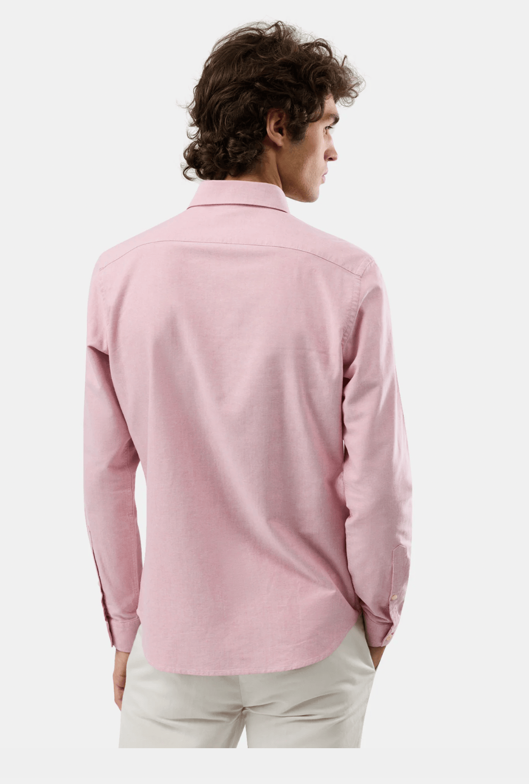 A man viewed from the back wearing a light pink long-sleeve button-down shirt and off-white trousers, set against a plain light gray studio background.