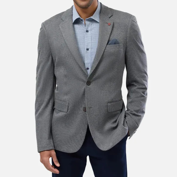 A man wearing a gray textured blazer over a light blue button-down shirt and dark navy trousers, standing against a light gray studio background.