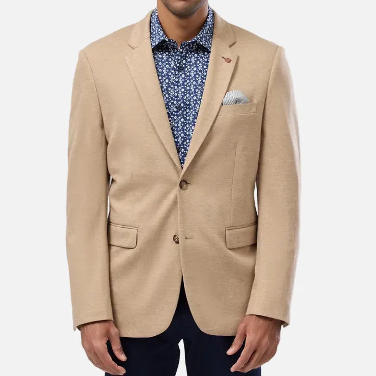 A man wearing a beige blazer over a blue patterned shirt, paired with dark trousers, standing against a light gray studio background.
