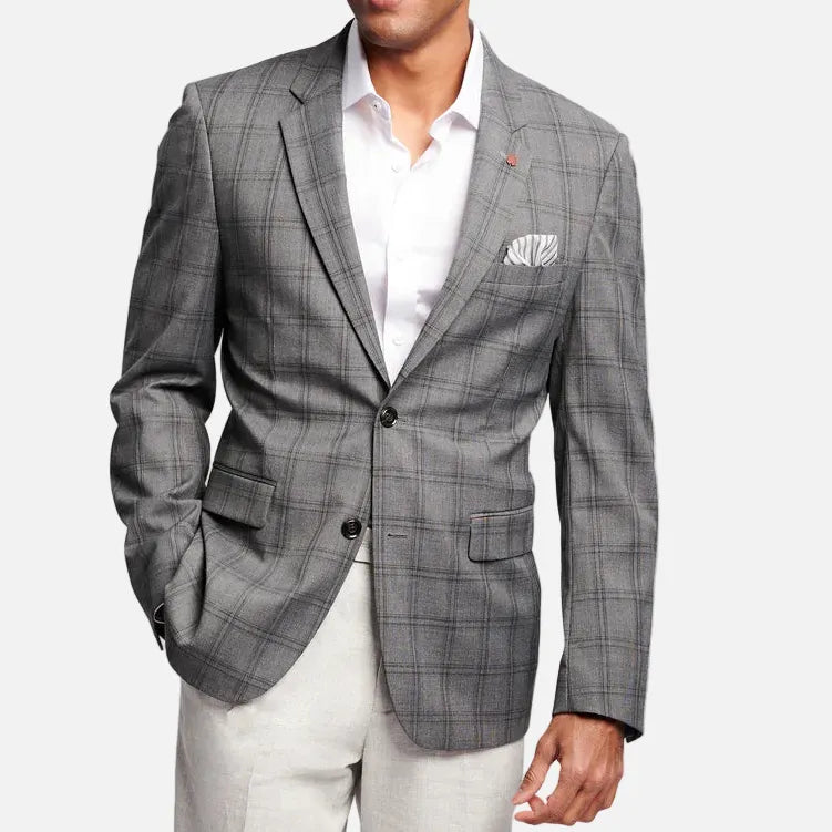 A man wearing a gray plaid blazer over a white dress shirt and light beige trousers, standing against a light gray studio background.
