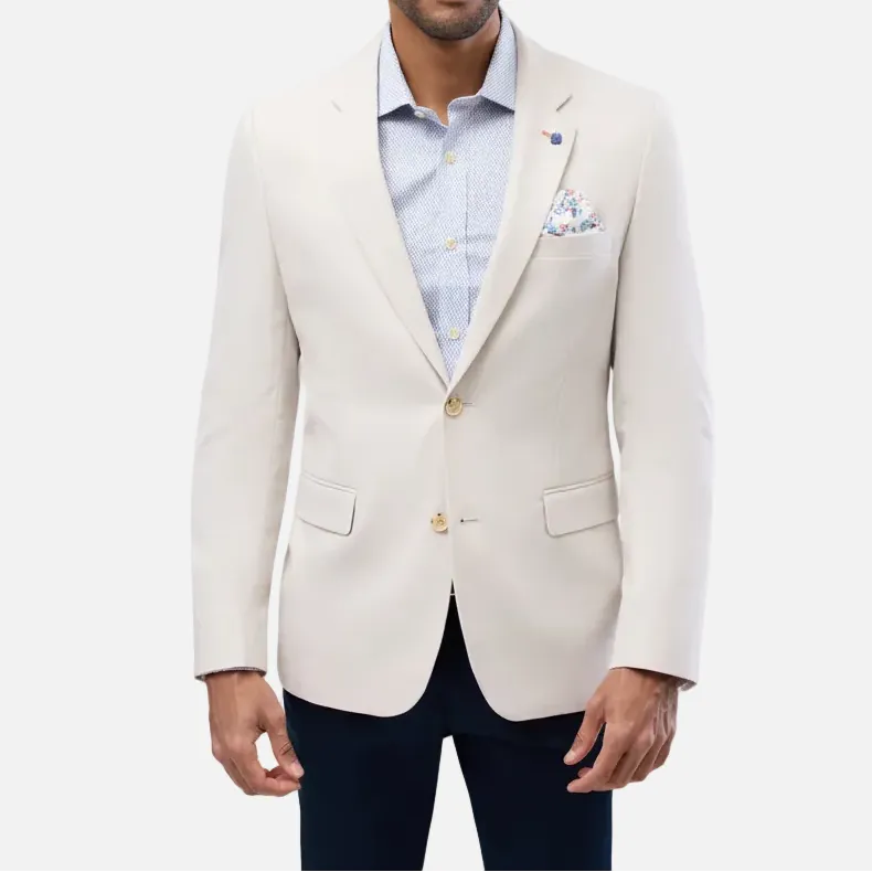 A man wearing a light cream-colored blazer over a light blue dress shirt and dark navy trousers, standing against a solid light gray background.